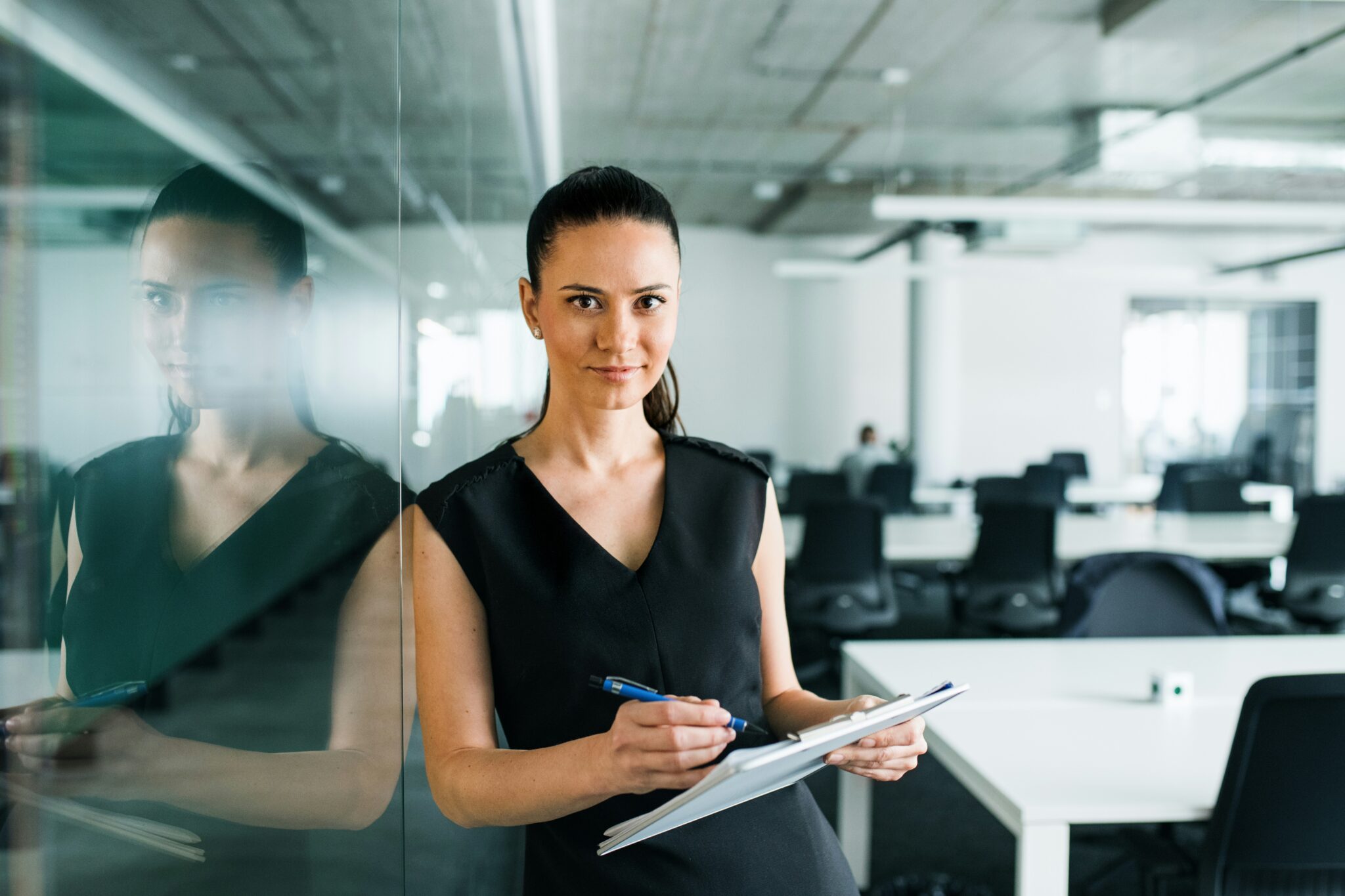 A confident woman in a black dress writing on a clipboard while standing in a modern office.