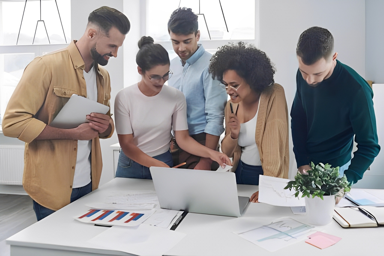 A diverse group of coworkers smiling and discussing something on a laptop screen in a bright workspace.